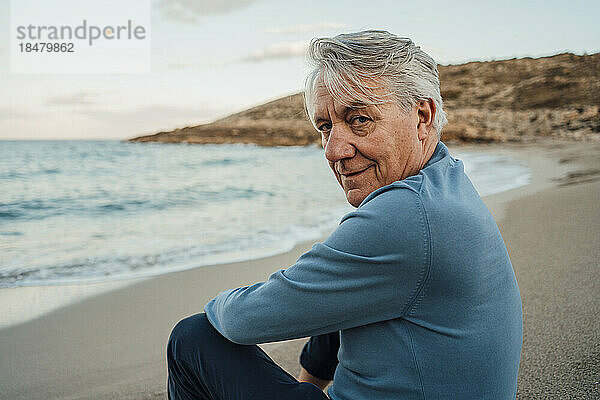 Lächelnder älterer Mann mit grauen Haaren sitzt am Strand
