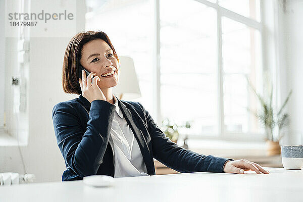 Glückliche Geschäftsfrau  die am Schreibtisch im Büro mit dem Mobiltelefon spricht