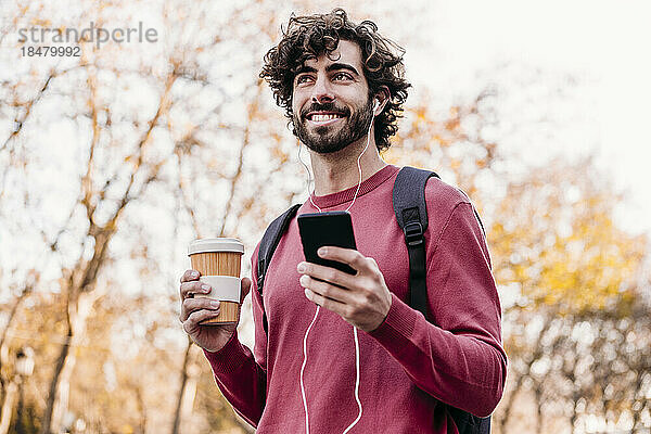 Lächelnder Mann mit Mobiltelefon und Kaffeetasse steht am Fußweg