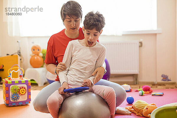 Physiotherapeutin und Mädchen mit Behinderung sitzen auf einem Fitnessball im Rehabilitationszentrum