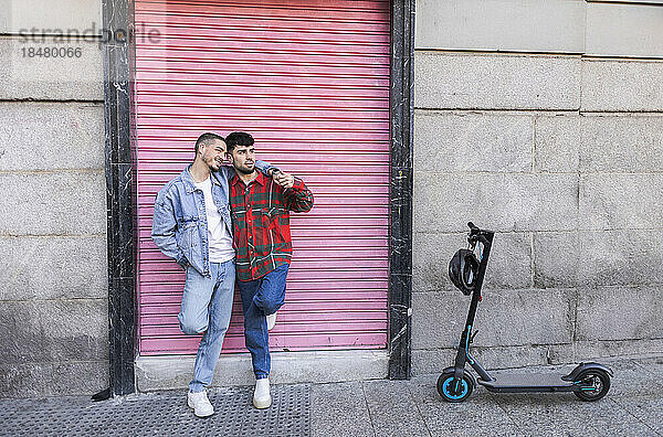 Glückliches schwules Paar  das mit einem Elektroroller vor dem Rollladen steht