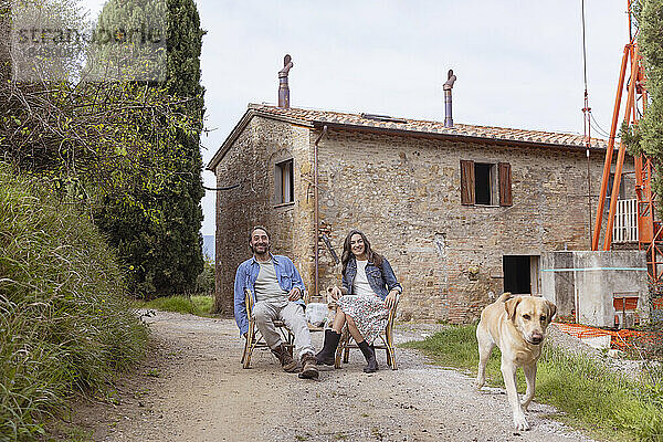 Glückliches älteres Paar sitzt mit Hund auf einem Stuhl vor dem Haus