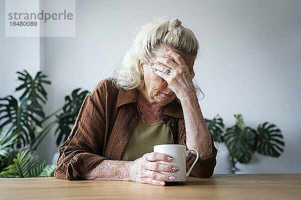 Deprimierte ältere Frau mit Tasse auf dem Tisch zu Hause