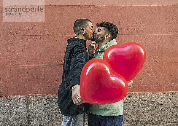 Affectionate gay couple kissing with red heart shape balloon in front of wall