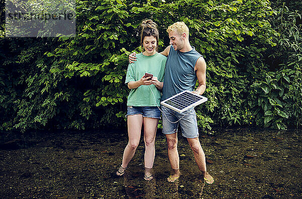 Glückliches Paar nutzt Smartphone und wird durch im Strom stehendes Solarpanel aufgeladen
