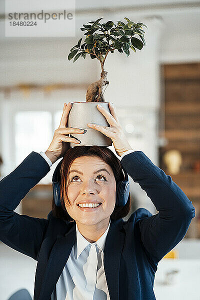 Glückliche junge Geschäftsfrau hält im Büro eine Topfpflanze auf dem Kopf