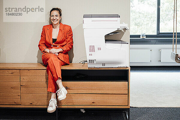 Lächelnde junge Geschäftsfrau sitzt am Drucker im Büro