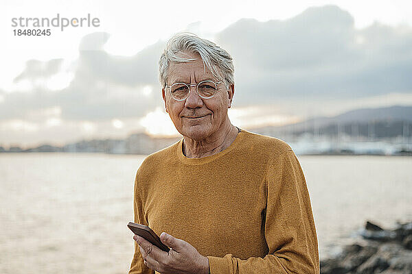 Lächelnder älterer Mann steht mit Mobiltelefon vor dem Meer