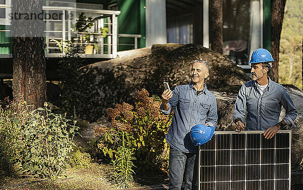 Photovoltaic technicians leaning on solar panel lookint at smartphone