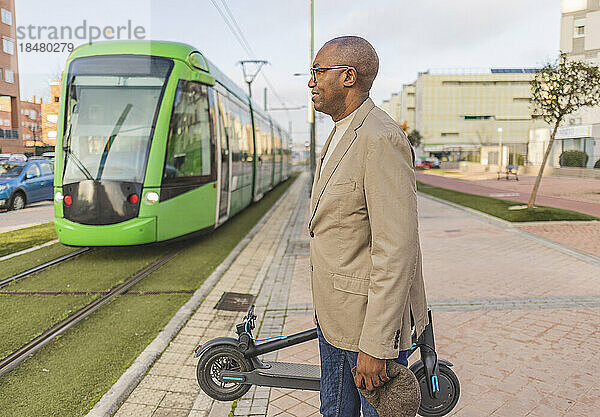 Geschäftsmann mit Elektroroller wartet auf Straßenbahn auf Fußweg