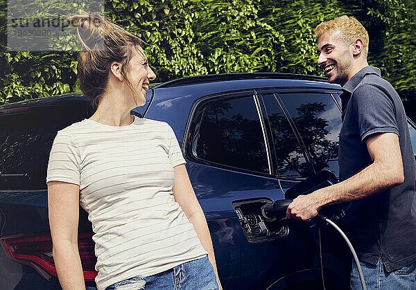 Glückliche Frau spricht mit ihrem Freund  der ein Elektroauto lädt