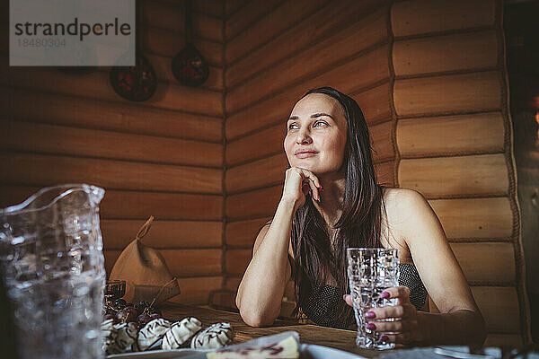 Lächelnde Frau mit Trinkglas  die in der Sauna nachdenkt