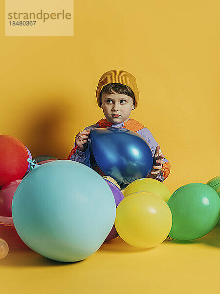 Junge sitzt mit bunten Luftballons vor gelbem Hintergrund