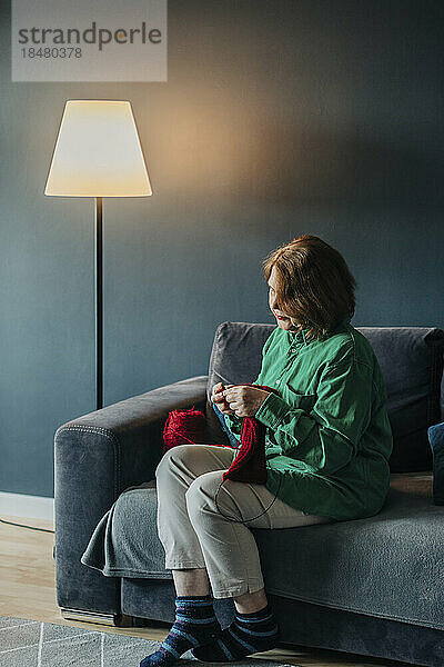 Ältere Frau strickt Pullover und sitzt zu Hause auf dem Sofa neben einer beleuchteten Lampe
