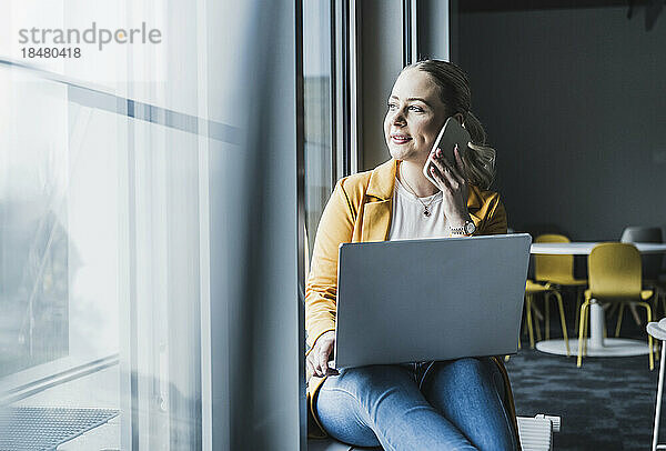 Lächelnde Geschäftsfrau  die am Fenster mit Laptop im Büro sitzt und am Smartphone spricht