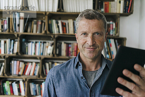 Lächelnder Mann mit Tablet-PC steht vor Bücherregal