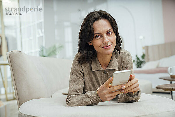 Lächelnde junge Frau mit Mobiltelefon  die zu Hause auf dem Sofa liegt