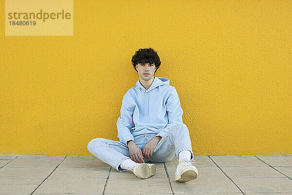 Selbstbewusster junger Mann sitzt auf Fußweg vor gelber Wand