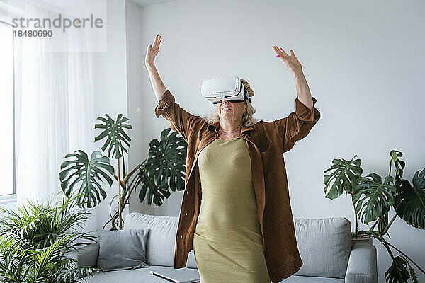 Sorglose Frau mit erhobenen Armen und Virtual-Reality-Headset steht zu Hause