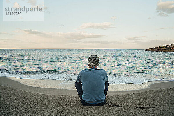 Älterer Mann sitzt auf Sand vor dem Meer am Strand