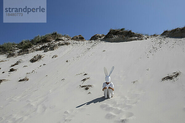 Frau mit Kaninchenmaske sitzt an sonnigem Tag auf Sanddüne