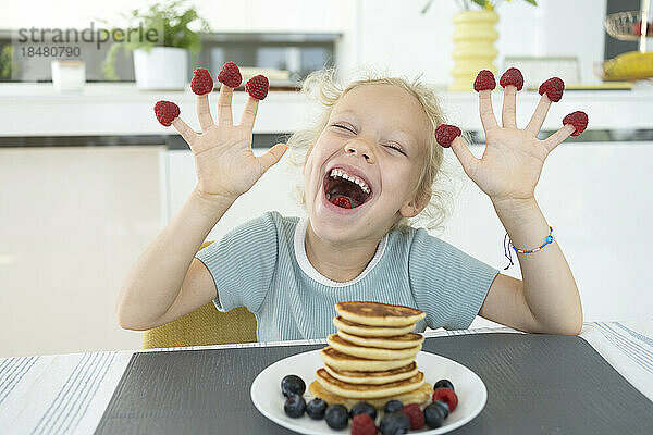 Playful girl with raspberries on fingers laughing at dining table at home