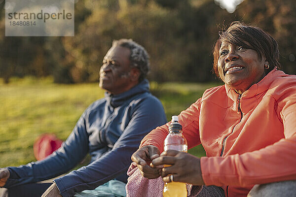 Smiling thoughtful woman with energy drink sitting by man in park