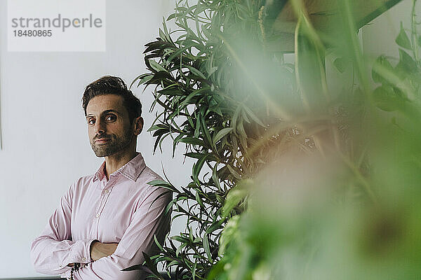 Selbstbewusster reifer Geschäftsmann steht mit verschränkten Armen neben Pflanzen im Büro