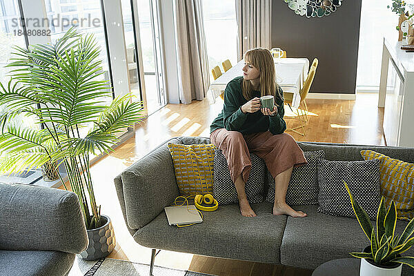 Nachdenkliche Frau mit Teetasse sitzt zu Hause auf dem Sofa