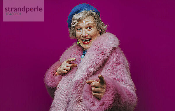 Glückliche ältere Frau mit rosa Pelzmantel gestikuliert vor farbigem Hintergrund