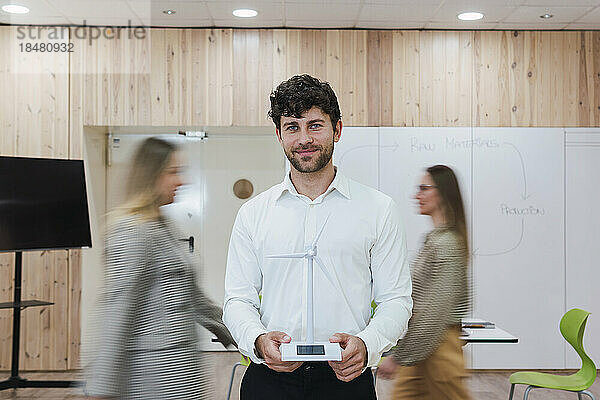 Businessman in office holding wind turbine model with businesswomen passing by