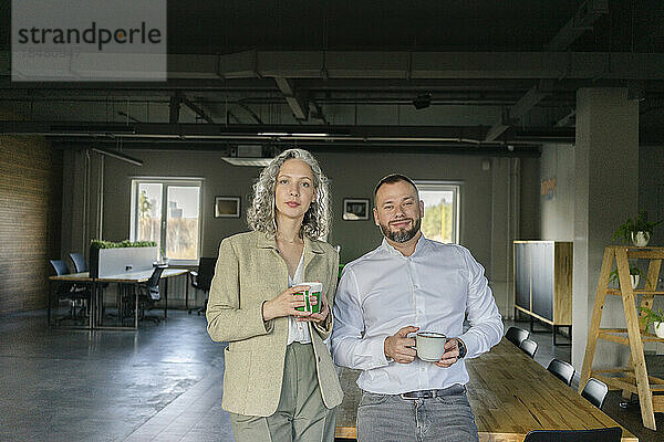 Portrait of confident businessman and businesswoman standing with coffee cups in office