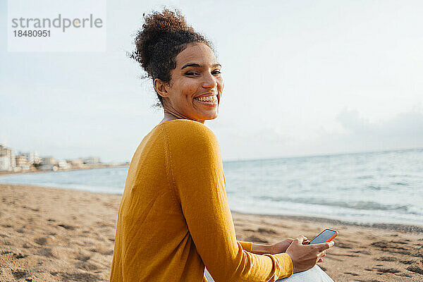 Glückliche Frau sitzt mit Handy am Strand