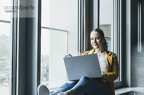 Lächelnde Geschäftsfrau mit Laptop sitzt am Fenster im Büro
