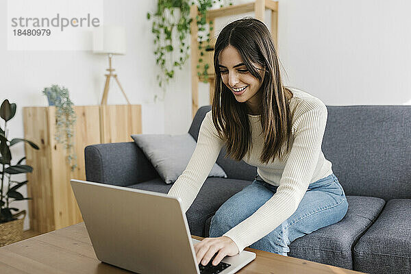 Glückliche junge Frau  die zu Hause auf dem Sofa sitzt und einen Laptop benutzt