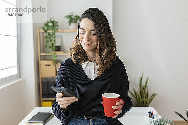 Lächelnde Geschäftsfrau mit Kaffeetasse und Smartphone im Büro