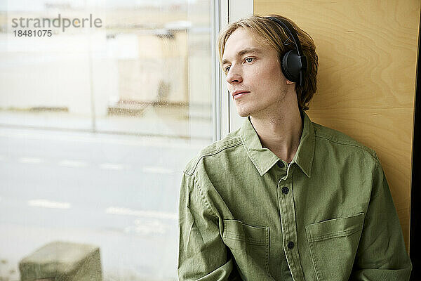 Junger Mann schaut aus dem Fenster und trägt Bluetooth-Kopfhörer