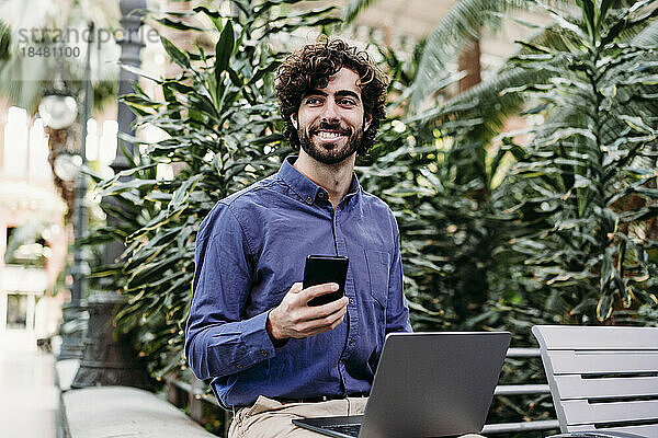 Lächelnder Geschäftsmann mit Laptop und Smartphone sitzt vor Pflanzen