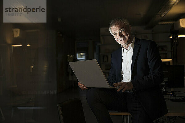 Lächelnder Geschäftsmann mit Laptop sitzt im Büro