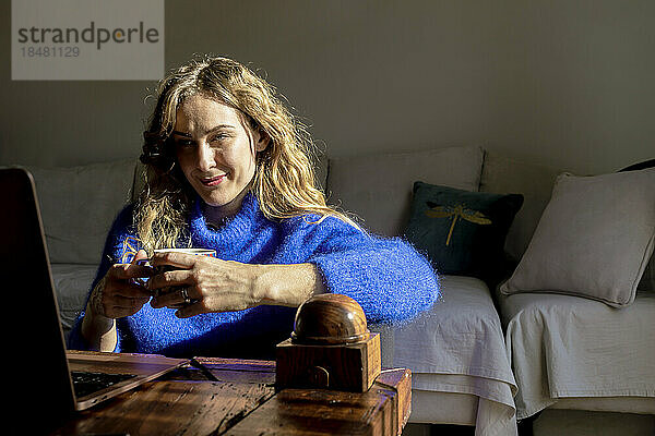 Lächelnde Frau mit Teetasse und Blick auf Laptop im Wohnzimmer
