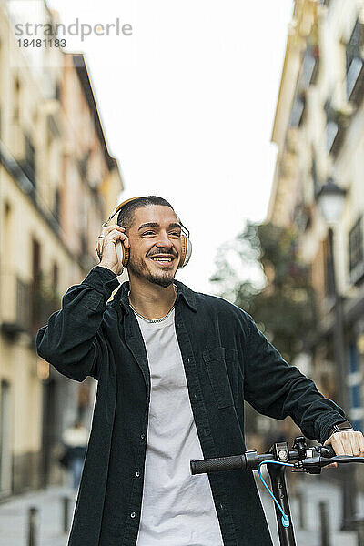 Glücklicher Mann mit kabellosen Kopfhörern  der den Griff eines elektrischen Tretrollers auf der Straße hält