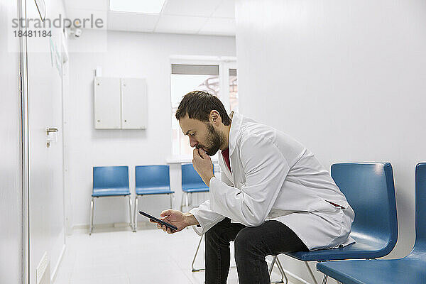 Besorgter Arzt sitzt mit Smartphone auf Stuhl im Flur