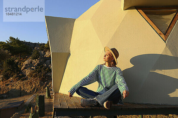 Sonnenlicht fällt auf eine Frau  die bei Sonnenuntergang mit gekreuzten Beinen in der Nähe eines nachhaltigen Gebäudes sitzt