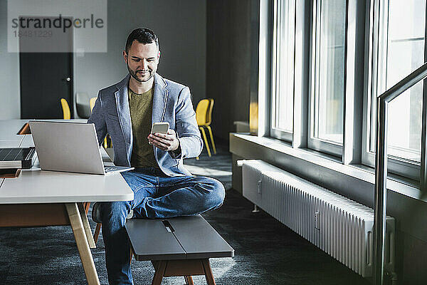 Geschäftsmann sitzt mit Smartphone auf Bank und Laptop am Schreibtisch im Büro