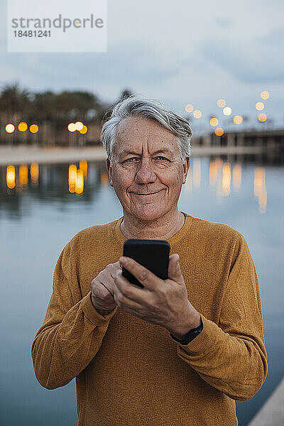 Smiling senior man standing with smart phone at sunset