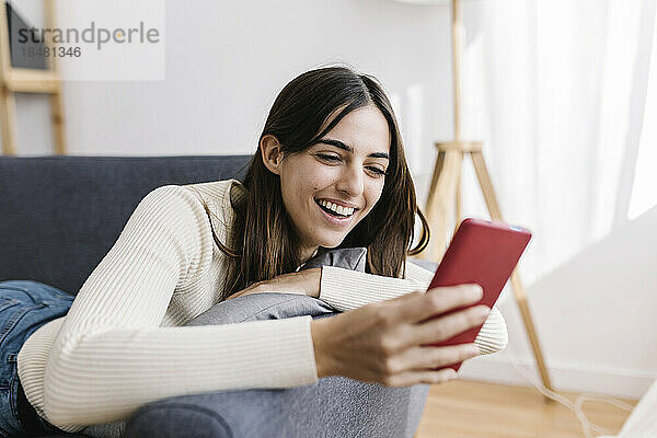 Glückliche junge Frau  die zu Hause auf dem Sofa liegt und ihr Smartphone benutzt