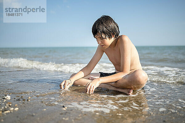 Boy playing in wet sand sitting cross-legged on shore at beach