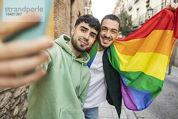 Glückliches schwules Paar mit Regenbogenfahne  das auf dem Fußweg ein Selfie mit dem Smartphone macht