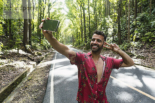 Glücklicher Mann zeigt eine Geste des Friedenszeichens und macht ein Selfie vor Bäumen