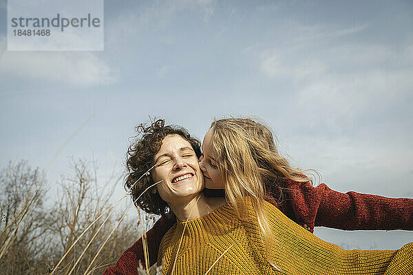 Girl with blond hair kissing mother in front of sky